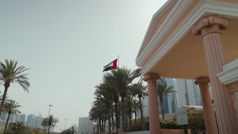 Vista-Panorámica-De-La-Entrada-De-La-Universidad-Americana-De-Dubái,-Con-Su-Edificio,-Las-Palmeras-Que-Lo-Rodean,-La-Bandera-De-Los-Emiratos-Árabes-Unidos-Y-La-Ciudad-De-Los-Medios-Y-El-área-Del-Puerto-Deportivo.