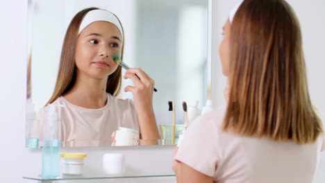 beauty,-skin-care-and-people-concept--teenage-girl-with-brush-applying-clay-mask-to-face-and-looking-in-mirror-at-bathroom