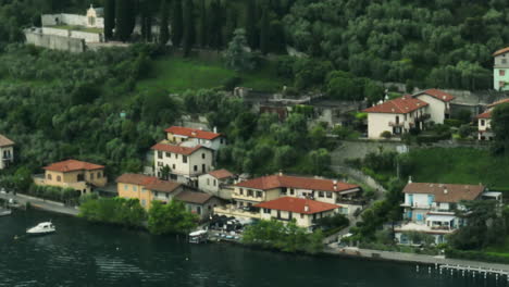 Vista-Panorámica-De-Un-Encantador-Pueblo-Junto-Al-Lago-Con-Barcos-Y-Exuberante-Vegetación-En-El-Lago-Iseo,-Italia