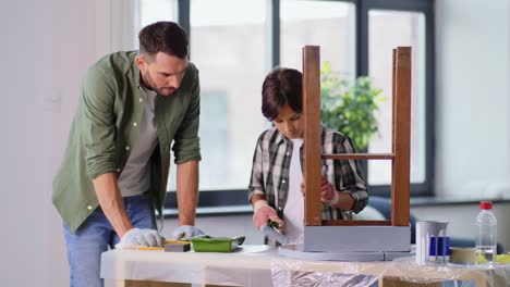 Concepto-De-Renovación,-Bricolaje-Y-Mejoras-Para-El-Hogar-Padre-E-Hijo-En-Guantes-Con-Rodillo-De-Pintura-Pintando-Una-Vieja-Mesa-De-Madera-En-Color-Gris-En-Casa