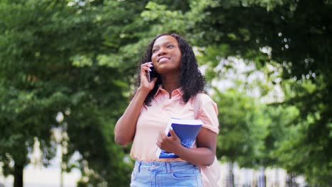 Concepto-De-Tecnología,-Educación-Y-Personas-Feliz-Y-Sonriente-Estudiante-Afroamericana-Con-Libros-De-Texto-Llamando-A-Un-Teléfono-Inteligente-En-La-Ciudad