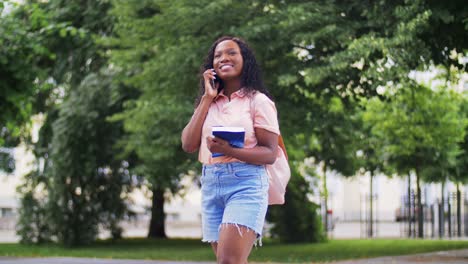 Concepto-De-Tecnología,-Educación-Y-Personas-Feliz-Y-Sonriente-Estudiante-Afroamericana-Con-Libros-De-Texto-Llamando-A-Un-Teléfono-Inteligente-En-La-Ciudad