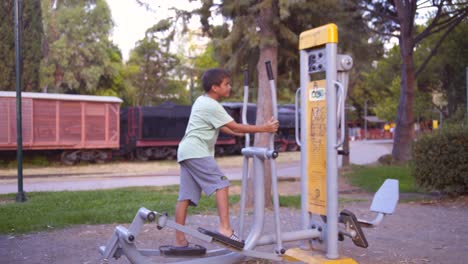 Wide,-side-view,-static-shot-of-8-yers-caucasian-boy,-entering-the-shot,-working-out-on-a-glide-gym-equipment,-located-at-Kalamata-railway-museum-park,-Greece