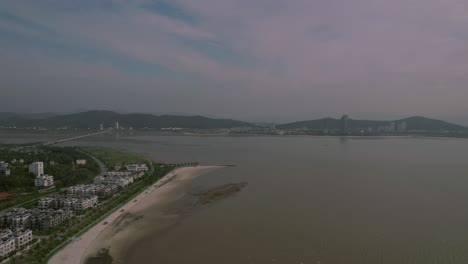 Aerial-coastal-scene-featuring-a-beachfront-lined-with-buildings,-a-sandy-beach,-and-mountains-in-the-distance-under-a-tranquil-sky,-capturing-nature's-serenity