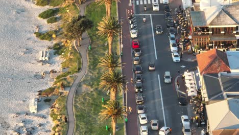 Vuelo-De-Un-Dron-Sobre-Un-Paseo-Marítimo-En-La-Playa-De-Camps-Bay-En-Ciudad-Del-Cabo,-Sudáfrica:-Coches-Circulando-Por-La-Carretera-Junto-A-Tiendas-Y-Gente-Local:-Palmeras-Y-Playa-Junto-A-La-Carretera