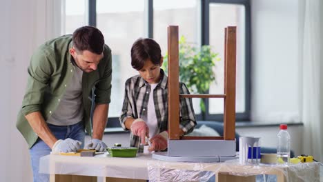 Concepto-De-Renovación,-Bricolaje-Y-Mejoras-Para-El-Hogar-Padre-E-Hijo-En-Guantes-Con-Rodillo-De-Pintura-Pintando-Una-Vieja-Mesa-De-Madera-En-Color-Gris-En-Casa