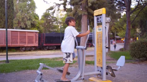 Wide,-static-side-view-of-caucasian-10-years-old-boy,-exercising-on-public-gym-equipment-,-at-Kalamata-railway-museum-park,-Greece