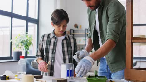 Reparatur-,-Heimwerken--Und-Heimwerkerkonzept-Vater-Und-Sohn-In-Schutzhandschuhen-Rühren-Graue-Farbe-In-Der-Dose-Um,-Um-Alten-Holztisch-Zu-Hause-Zu-Bemalen