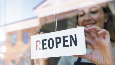 Concepto-De-Pequeña-Empresa,-Reapertura-Y-Servicio-Mujer-Sonriente-Feliz-Colgando-Pancarta-De-Reapertura-En-El-Vidrio-De-La-Ventana-O-Puerta