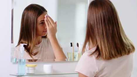 beauty,-hygiene-and-people-concept--teenage-girl-applying-moisturizer-to-her-face-and-looking-in-mirror-at-bathroom