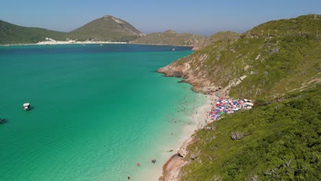 Un-Dron-Sobrevuela-Prainhas-Do-Atalaia-En-Arraial-Do-Cabo,-Una-Impresionante-Playa-De-Arena-Blanca-Con-Aguas-Cristalinas-Y-Pintorescas-Montañas-Insulares-Bajo-Un-Cielo-Azul-Claro.