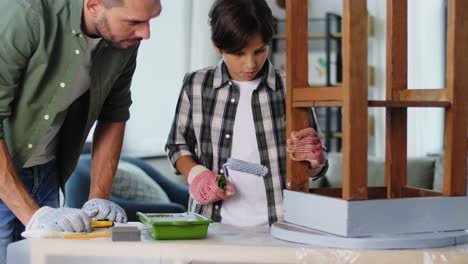 Concepto-De-Renovación,-Bricolaje-Y-Mejoras-Para-El-Hogar-Padre-E-Hijo-En-Guantes-Con-Rodillo-De-Pintura-Pintando-Una-Vieja-Mesa-De-Madera-En-Color-Gris-En-Casa