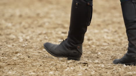 Close-up-of-a-person's-boots-walking-on-a-sandy-arena-surface