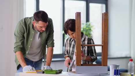 renovation,-diy-and-home-improvement-concept--father-and-son-in-gloves-with-paint-roller-painting-old-wooden-table-in-grey-color-at-home