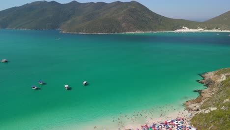 Drone-panning-left-view-over-Arraial-do-Cabo's-pristine-beaches,-capturing-the-stunning-blue-waters,-the-serene-island's-coastline,-and-the-majestic-mountains-of-an-island-in-the-background