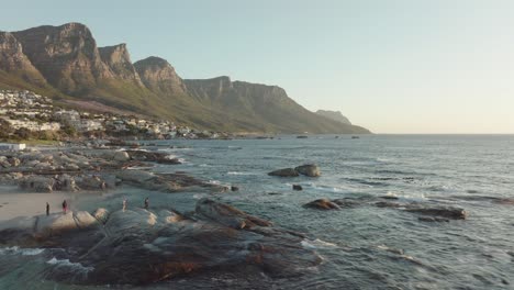 Drohne-Fliegt-Langsam-Rückwärts-Am-Camps-Bay-Beach-In-Kapstadt,-Südafrika---Leute-Sind-Am-Felsigen-Strand-Und-Genießen-Den-Sonnenuntergang---Blick-Auf-Die-Tafelbergfelsen-Im-Meer