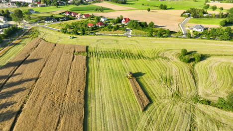 Vista-Aérea-De-Una-Cosechadora-Cortando-Un-Campo,-Con-Casas-Y-Una-Carretera-Visibles-Al-Fondo