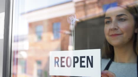 Kleines-Unternehmen,-Wiedereröffnung-Und-Service-Konzept-Glücklich-Lächelnde-Frau-Hängt-Wiedereröffnungsbanner-An-Fenster-Oder-Türglas