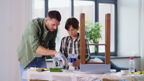 renovation,-diy-and-home-improvement-concept--father-and-son-in-gloves-with-paint-roller-painting-old-wooden-table-in-grey-color-at-home