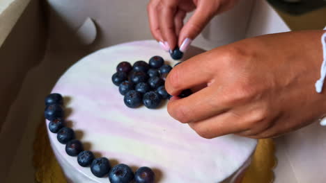 Close-up-view-of-woman's-hands-with-pink-nails-placing-blueberry-fruit-on-top-of-vanilla-cake-in-cardboard-box-celebration-dessert-decoration-icing-birthday-wedding