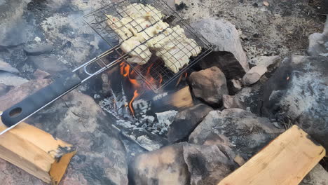 Cooking-meat-under-flaming-hot-charcoal,-outdoor-pit-fire-picnic-food,-smoke-coming-out