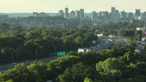 Nashville-city-skyline-with-lush-green-forest-and-busy-highway-at-sunset,-aerial-view