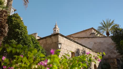 View-captures-an-elegant-stone-structure-surrounded-by-lush-green-trees-and-vibrant-flowers,-with-a-clear-blue-sky-and-a-tower-in-the-background-adding-to-its-charm