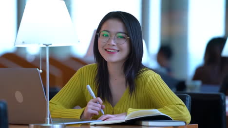 Portrait-of-happy-joyful-pretty-female-student-busy-hard-working-sitting-against-background-of-bookshelves-in-university-library-holding-laptop-and-backpack-looking-to-the-side,-smiling-pleasant