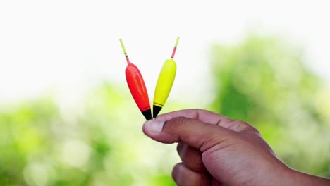 Bobber-De-Pesca-Colorido-Sostenido-Con-La-Mano-Que-Se-Muestra-En-La-Cámara-Sobre-Un-Fondo-Borroso