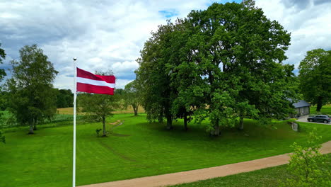 Bandera-De-Letonia-Ondeando-En-Una-Finca-Rural-Verde,-Vista-Aérea