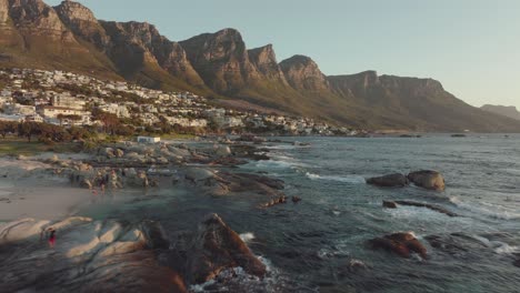 Drohne-Fliegt-Knapp-über-Dem-Meer-Am-Camps-Bay-Beach-In-Kapstadt,-Südafrika-–-Welliges-Meer,-Viele-Häuser-Auf-Einem-Hügel-–-Drohne-Fliegt-Rückwärts-Mit-Blick-Auf-Die-Tafelbergfelsen-Im-Meer