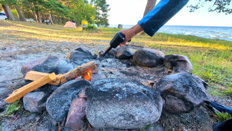 Shot-of-person-holding-electric-fire-blower-on-campsite-flames-wooden-smoky-logs-rocky-fire-pit-with-tents-beside-lake-sea-travel-camping-outdoors-wilderness