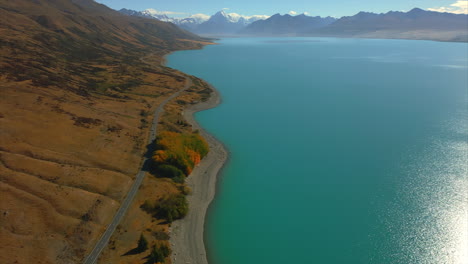 Lake-Pukaki-along-a-countryside-road---aerial-tilt-up-to-reveal-Mont-Cook,-Aoraki,-New-Zealand