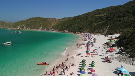 Stunning-aerial-drone-flyover-Atalaia-Point-Beach-,-Arraial-do-Cabo,-Cabo-Frio-capturing-a-white-sandy-beach,-crystal-blue-waters,-and-majestic-mountains-in-the-background