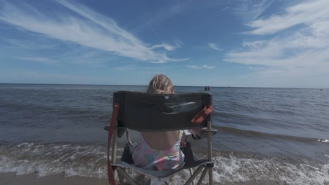 Girl-sitting-on-a-beach-chair,-facing-the-sea-under-a-blue-sky-adorned-with-wispy-clouds