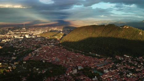 Vista-Aérea-Panorámica-De-La-Ciudad-De-Brasov-Al-Atardecer-Con-Nubes-Espectaculares-Y-Exuberantes-Colinas-Verdes