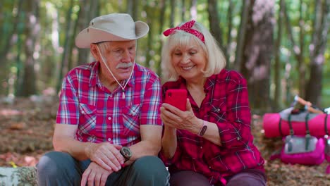 Attractive-senior-old-man-woman-backpacker-planning-trip-route-in-forest-on-mobile-phone-in-wood