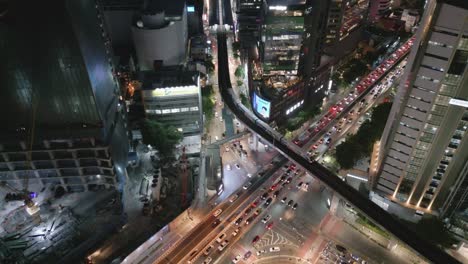 Vista-Aérea-De-La-Intersección-Del-Tráfico-Vial-Del-Metro-De-Tránsito-Rápido-O-MRT-Skytrain-Iluminada-Por-La-Noche