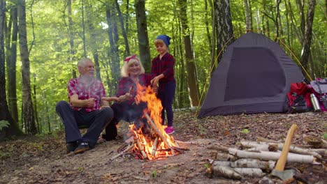 Senior-grandmother-grandfather-with-granddaughter-cooking-frying-sausages-over-campfire-in-wood