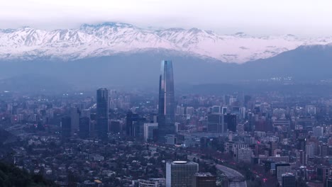 Aerial-drone-view-right-pan-of-snowy-Andes-mountains-and-Santiago-de-Chile-city-buildings-and-Costanera-Center-Mall-on-a-cloudy-winter-morning