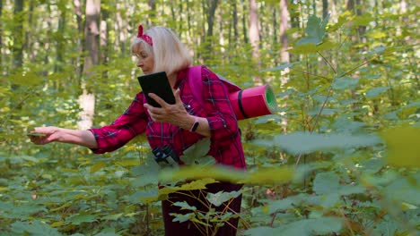 Active-senior-elderly-grandmother-adventurer-exploring-forest-trees,-plants-with-her-digital-tablet