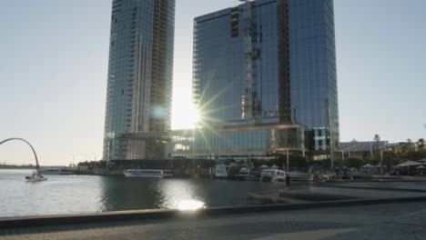 Public-transportation-boat-harbour-at-Elizabeth-Quay-during-golden-hour,-sunlight-rays-behind-tall-building,-Timelapse