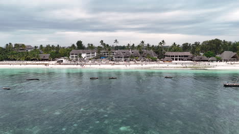 Aerial-approaching-shot-of-coral-in-water,-tourist-at-sandy-beach-and-luxury-resort-with-palm-trees-on-Zanzibar-Island,-Africa