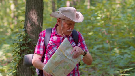 Senior-old-tourist-grandfather-man-lost-and-looking-at-map-and-compass-while-having-walk-in-wood