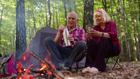 Senior-ältere-Großmutter-Großvater-Gießt-Getränk-Aus-Thermoskanne-Trinken-Tee-über-Lagerfeuer-In-Holz