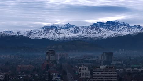 Aerial-drone-zoom-in-view-of-snowy-Andes-mountains-and-Santiago-de-Chile-city-buildings-on-a-cloudy-winter-morning