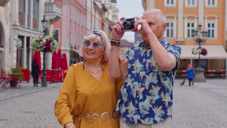 Elderly-stylish-tourists-man-and-woman-taking-photos-with-old-film-camera,-walking-along-city-street