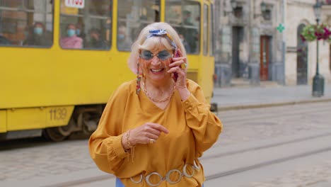 Senior-old-tourist-woman-in-stylish-clothes-talking-on-mobile-phone-while-walking-on-city-street