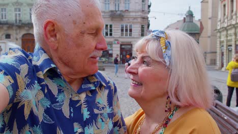 POV-shot-of-senior-tourists-couple-taking-selfie-in-summer-city-center-smiling,-looking-at-camera