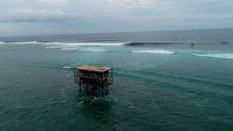 Judging-tower-structure-looks-out-to-waves-at-Cloudbreak-Fiji-with-surfers,-aerial-dolly